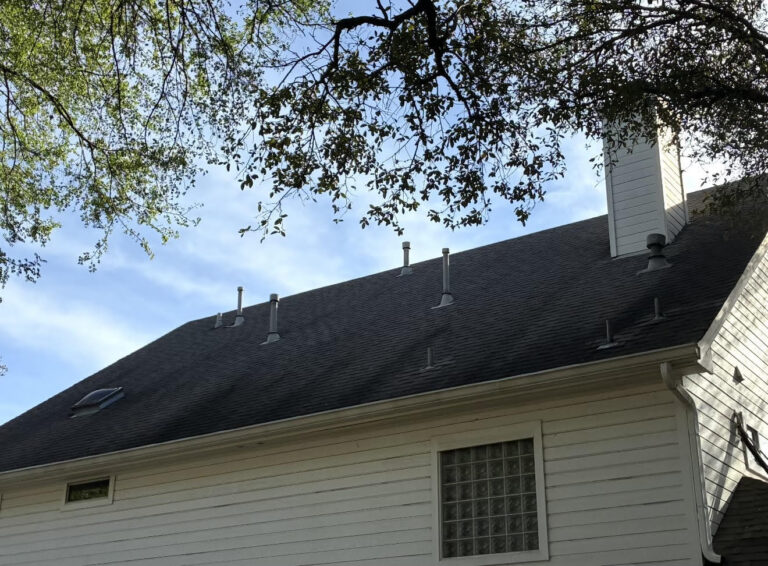 Before_1 - Driftwood Builders Roofing White house with sloped roof and multiple vent pipes, skylight, and tall chimney, viewed from below with tree branches overhead.