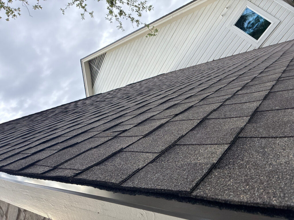 Close-up angled view of a dark shingled roof against a white siding dormer with a small rectangular window and vent.