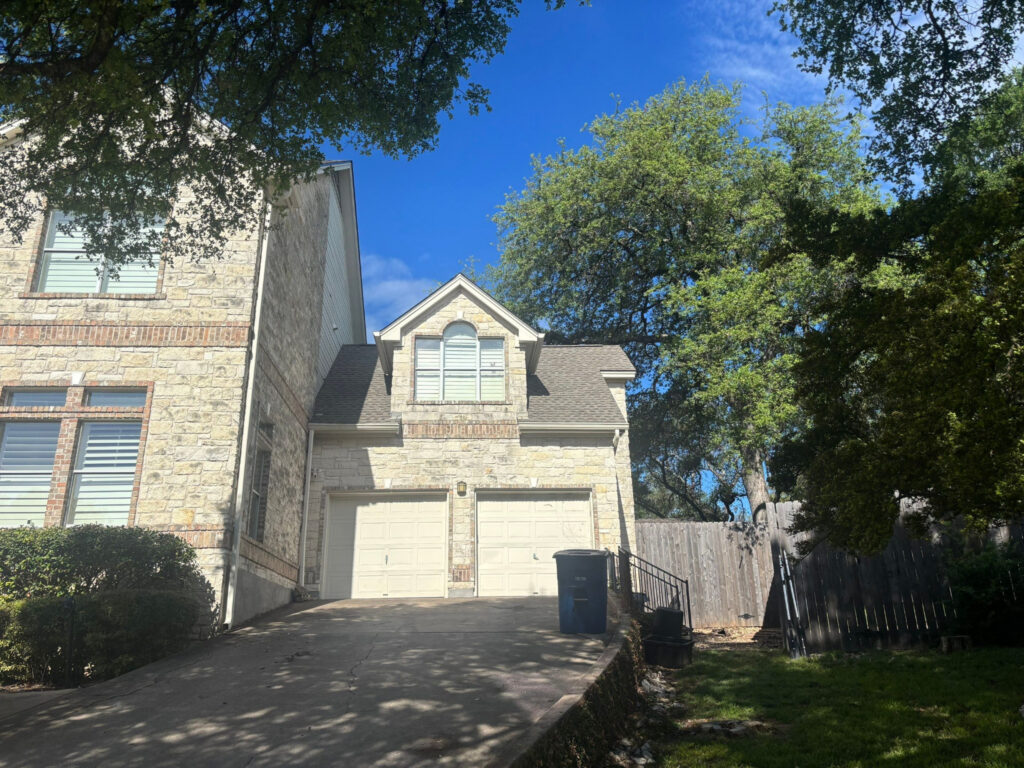 Two-car stone house with white garage doors, driveway, and a large tree canopy overhanging the front yard under a blue sky.
