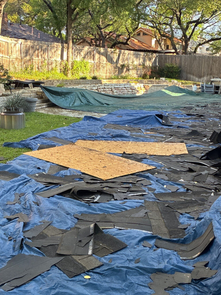 During_1 - Driftwood Builders Roofing Backyard renovation area with blue tarps and scattered black roofing shingles over a paved surface, a stone retaining wall, and a wooden fence in the background.