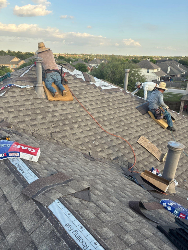 during_01 - Driftwood Builders Roofing Three construction workers install new asphalt shingles on a sloped suburban roof, kneeling on pads with tools nearby during daytime sun. Often wearing wide-brimmed hats for sun protection.