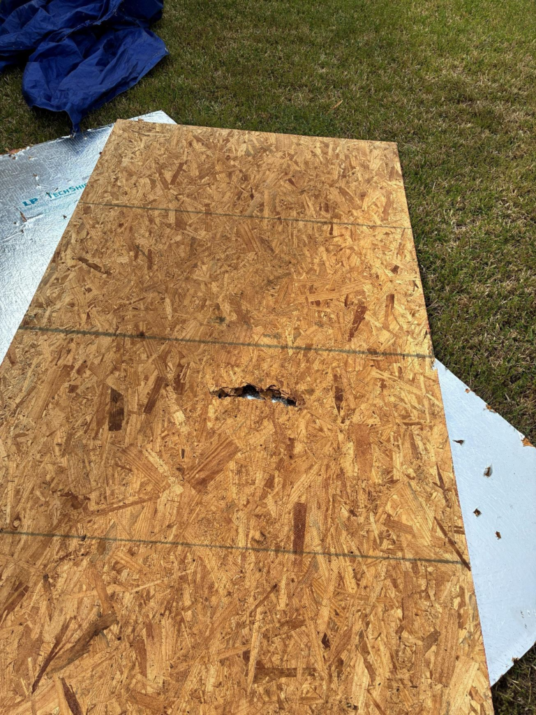 during_06 - Driftwood Builders Roofing OSB plywood sheet on grass with a small hole along a seam, nearby blue tarp partially visible. Likely construction site materials.