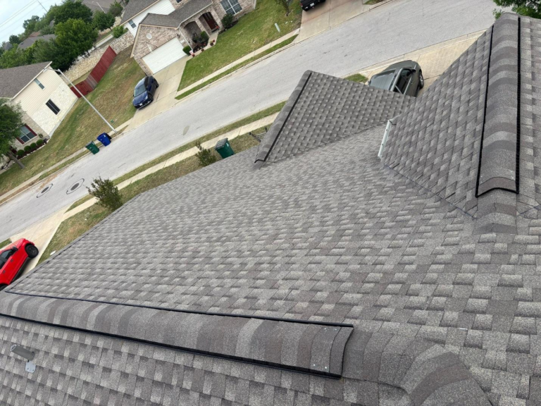 after_02 - Driftwood Builders Roofing Aerial view of a gray shingled roof with a vent pipe and dormer, overlooking a suburban street with parked cars and green lawns.