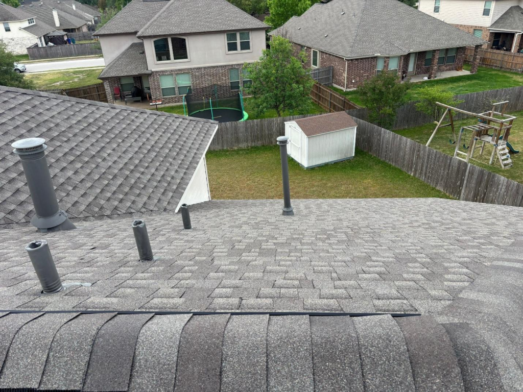 View from a gray shingled roof onto a suburban backyard with a trampoline, shed, and wooden fence enclosing grassy yards.