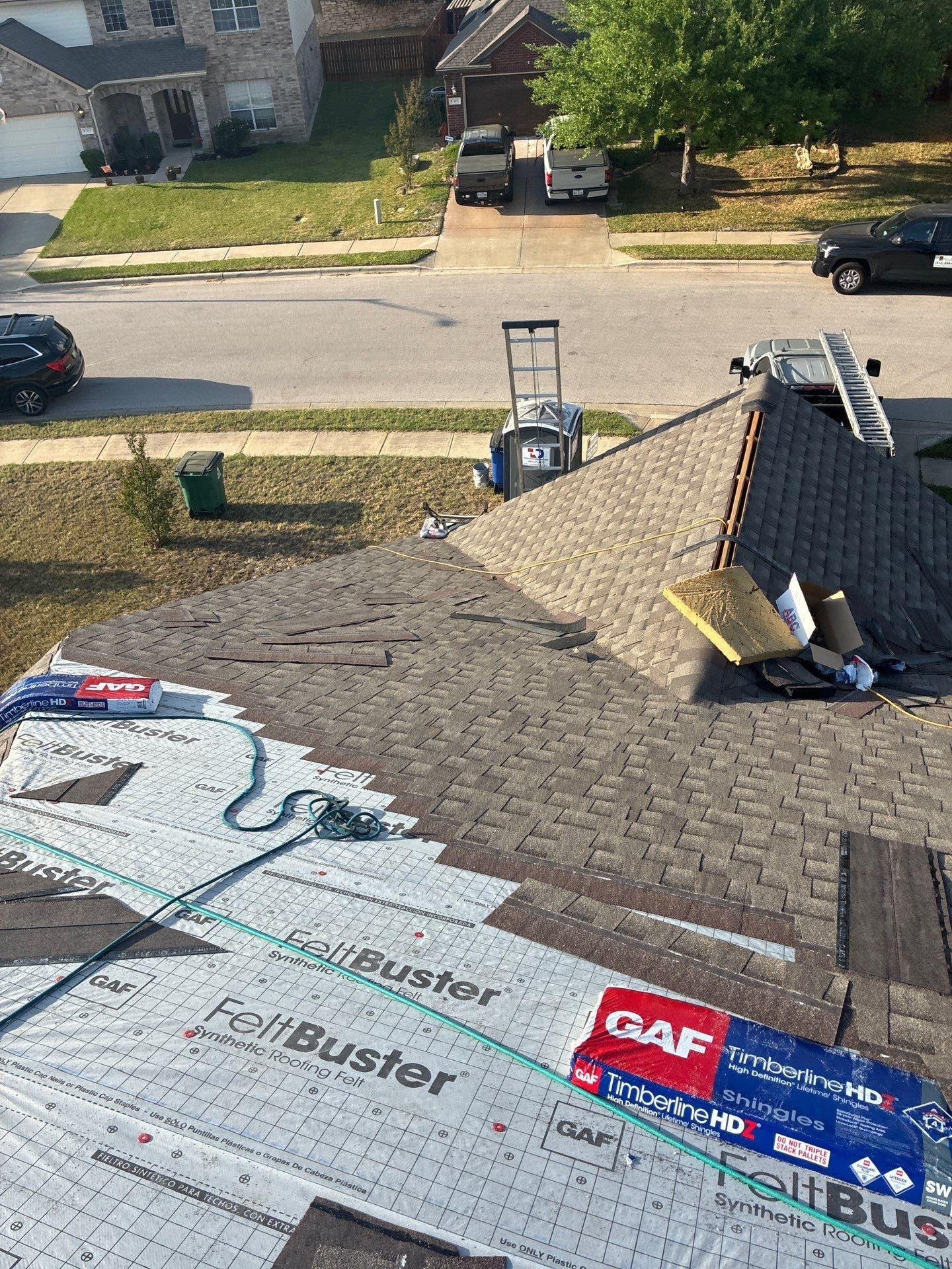 during_04 - Driftwood Builders Roofing - Driftwood Builders Roofing Rooftop under construction with FeltBuster roofing underlayment, a ladder, and bundles of shingles; a suburban street with parked cars below.