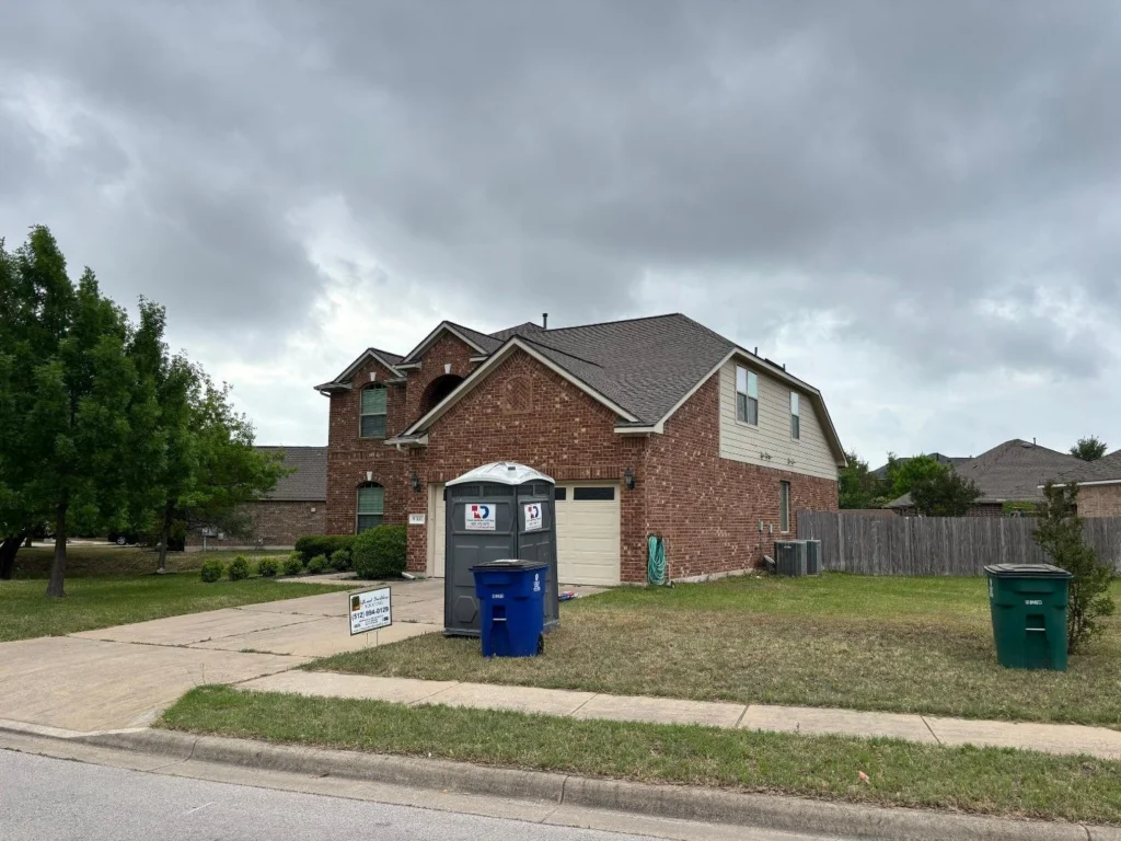 Two-story brick suburban house with attached garage and a portable toilet in the driveway, plus blue recycling and green trash bins nearby under a cloudy sky.