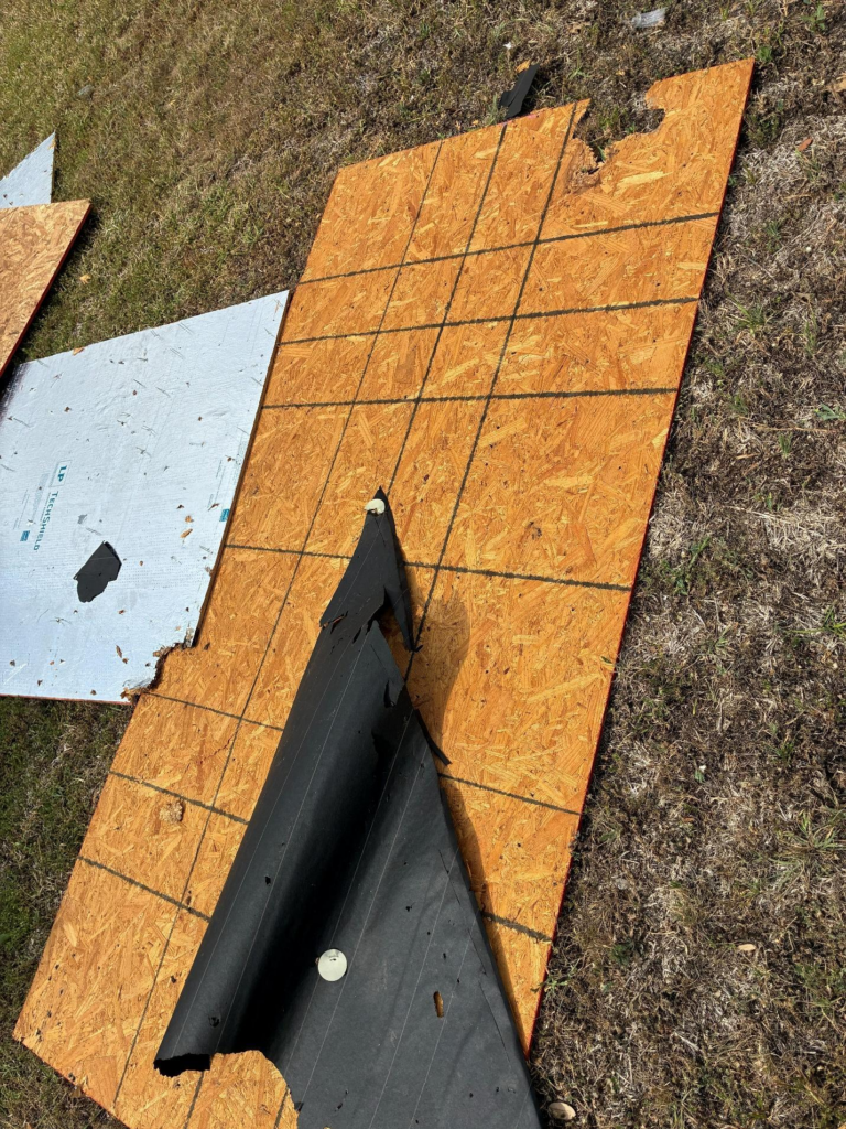 during_07 - Driftwood Builders Roofing Outdoor scene with large sheets of oriented strand board (OSB) plywood and torn black roofing membrane lying on dry grass; a gray insulated panel is visible on the left. Debris scattered across a construction site.