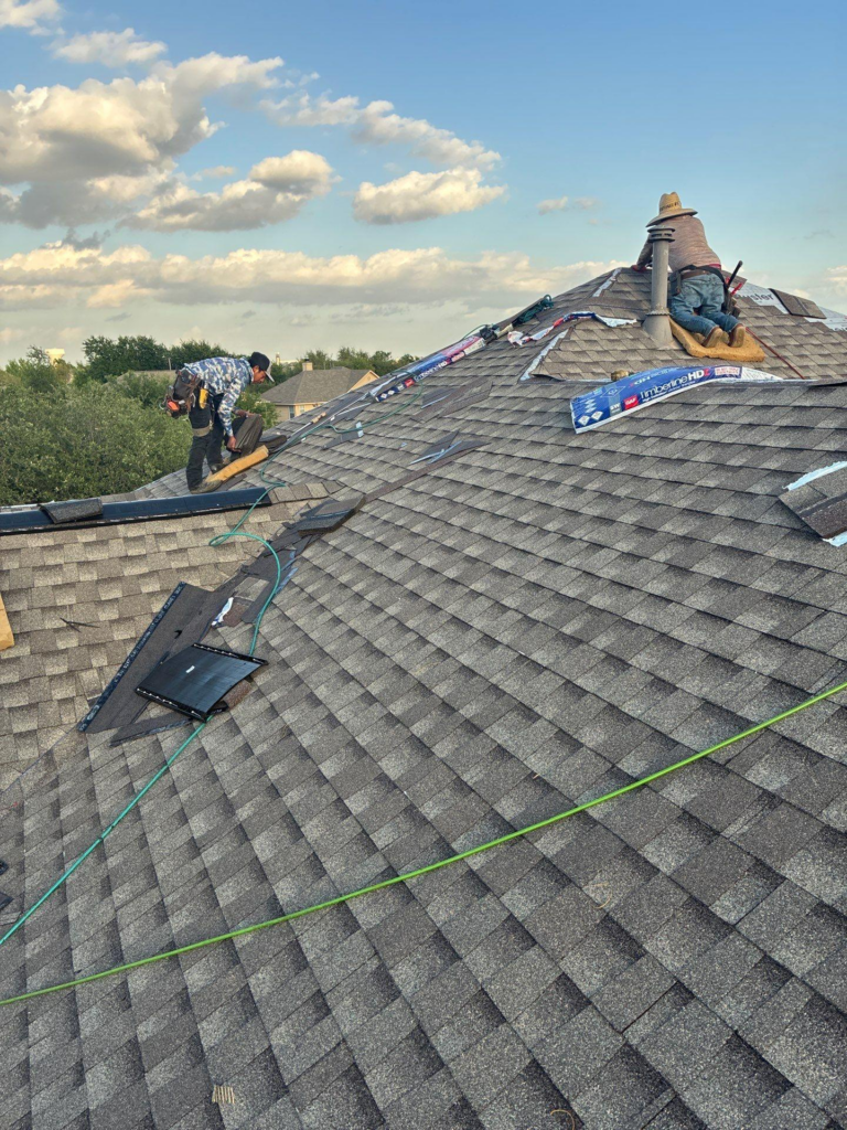 during_02 - Driftwood Builders Roofing Two workers on a sloped roof repairing shingles, with tools and a vent pipe nearby under a blue sky with clouds