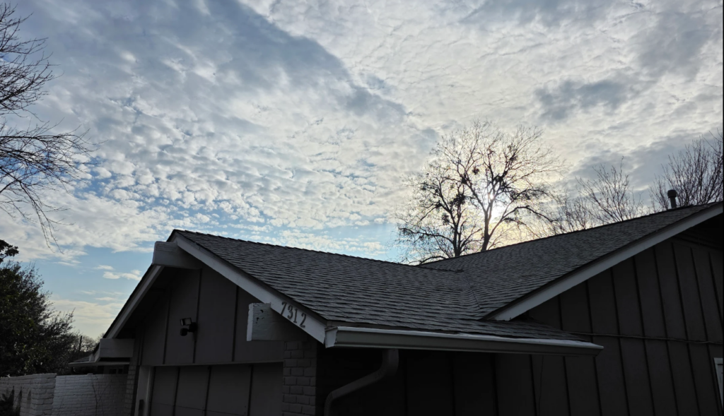 Low-angle view of a suburban house with gray siding and a sloped roof, sun peeking through a bare tree behind the roof under a cloudy sky.