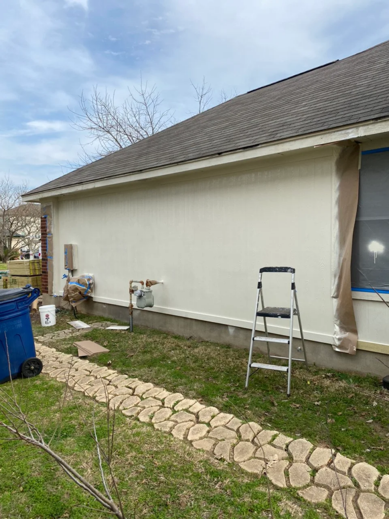 Leander_During_2 1 - Driftwood Builders Roofing Exterior side of a house under renovation with a ladder, painter's tarp by a window, and a blue recycling bin; supplies and cardboard on the ground nearby.