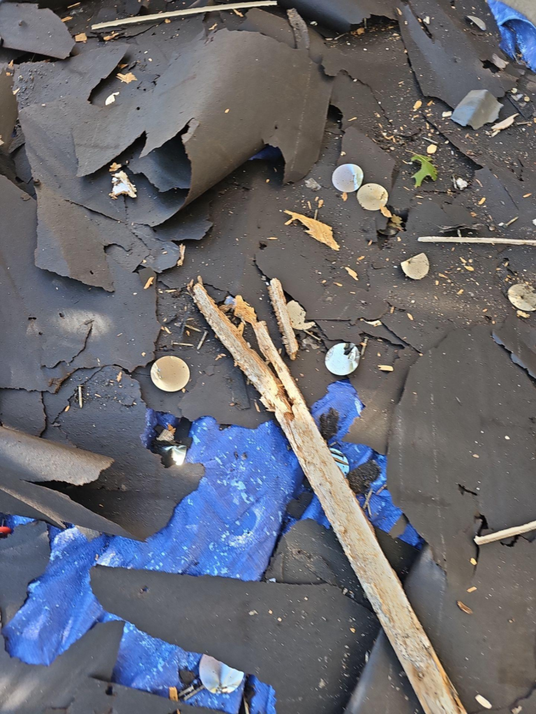during_03_debris - Driftwood Builders Roofing Pile of torn black roofing shingles and debris with blue tarp showing beneath, plus small wooden sticks and round metal washers scattered about