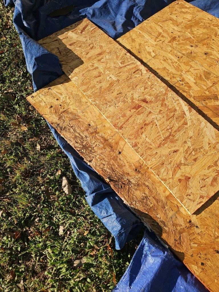 during_01_decking_removed - Driftwood Builders Roofing OSB plywood sheets laid on a blue tarpaulin outdoors, resting on grass and leaves nearby.