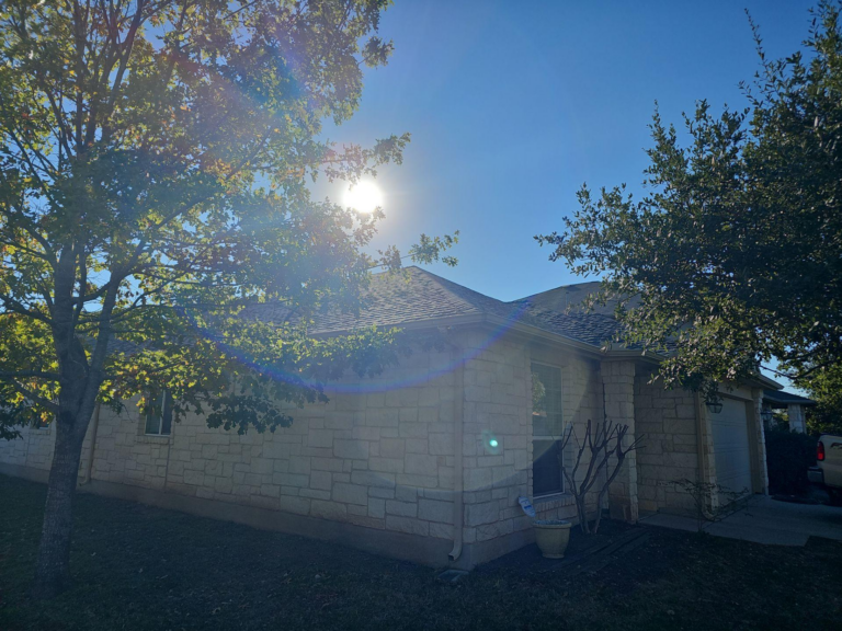 after_01_front_completed - Driftwood Builders Roofing Front view of a beige stone house with a sloped roof, framed by two trees and a sun glare in the sky.
