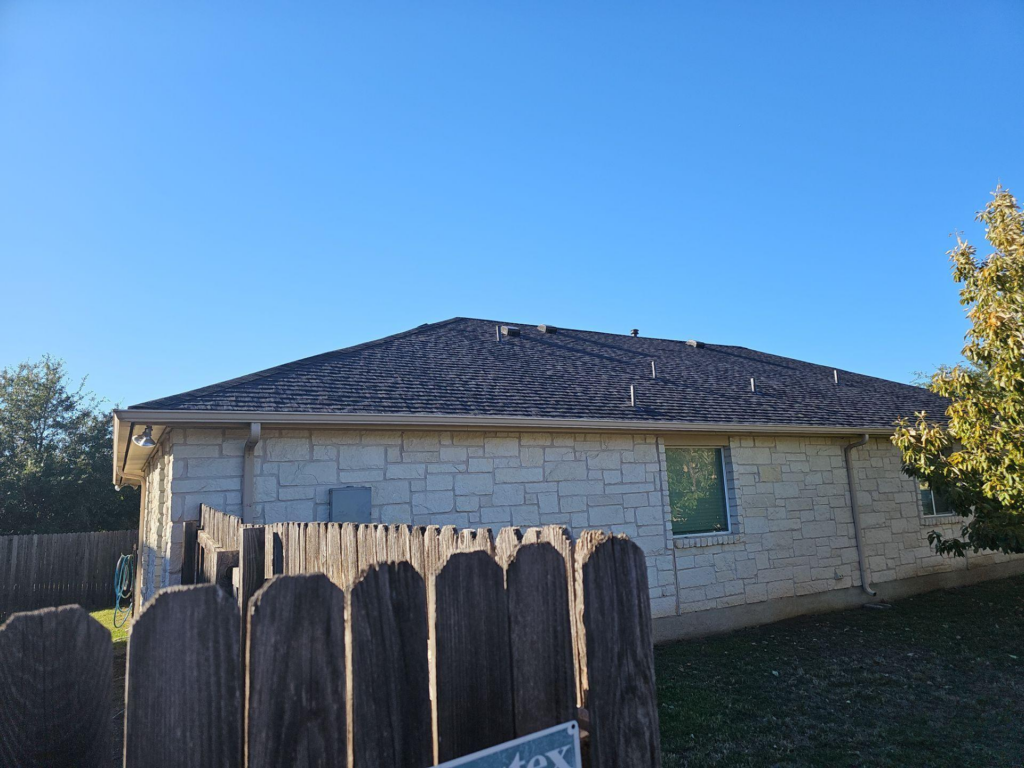 Backyard view of a stone house with a dark roof, a wooden fence in the foreground, and a clear blue sky on a sunny day.