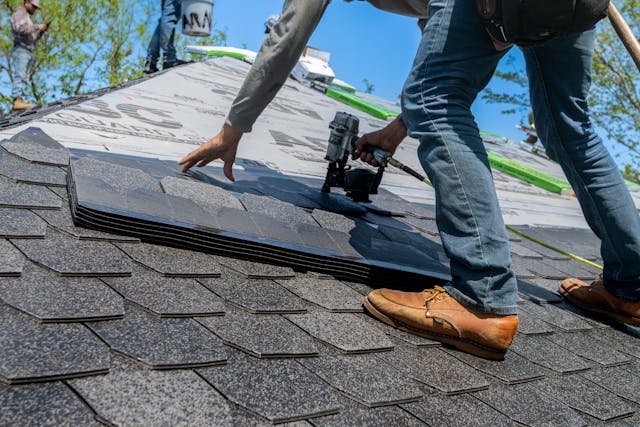 Roofer on a sloped roof places dark asphalt shingles, using a nail gun nearby.