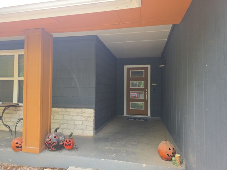 Front porch of a house with blue siding and an orange support beam, brown door with glass panels, and several jack-o'-lanterns on the steps outside.