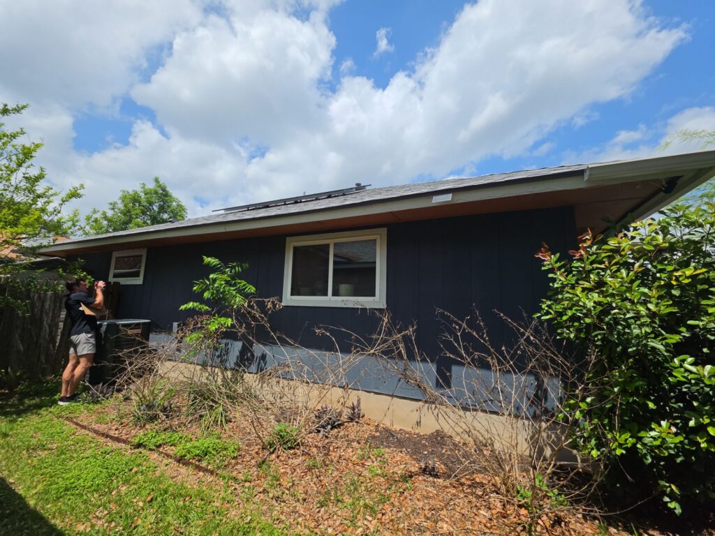 Man wearing black shirt and shorts taking a photo of a blue single-story house with green shrubs and a cloudy sky above.