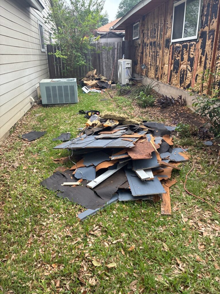 Backyard scene with torn wall siding and a large pile of debris (wood, metal panels) next to an outdoor air conditioner. An exposing fence and plants surround the area.