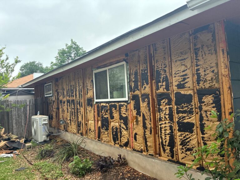 Exterior of a house undergoing renovation with insulation exposed between studs and a window visible on the wall, and an outdoor HVAC unit nearby.