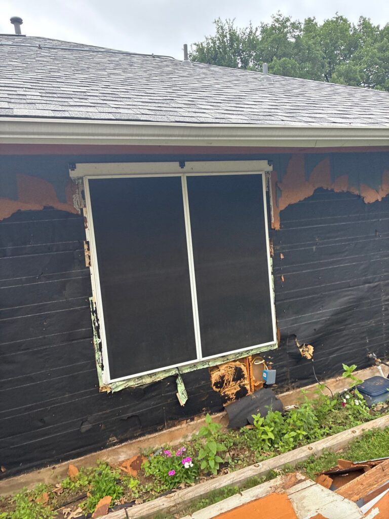 Sliding glass door on a house under renovation; black siding peeling away to reveal orange underlayment and scattered debris in the yard.