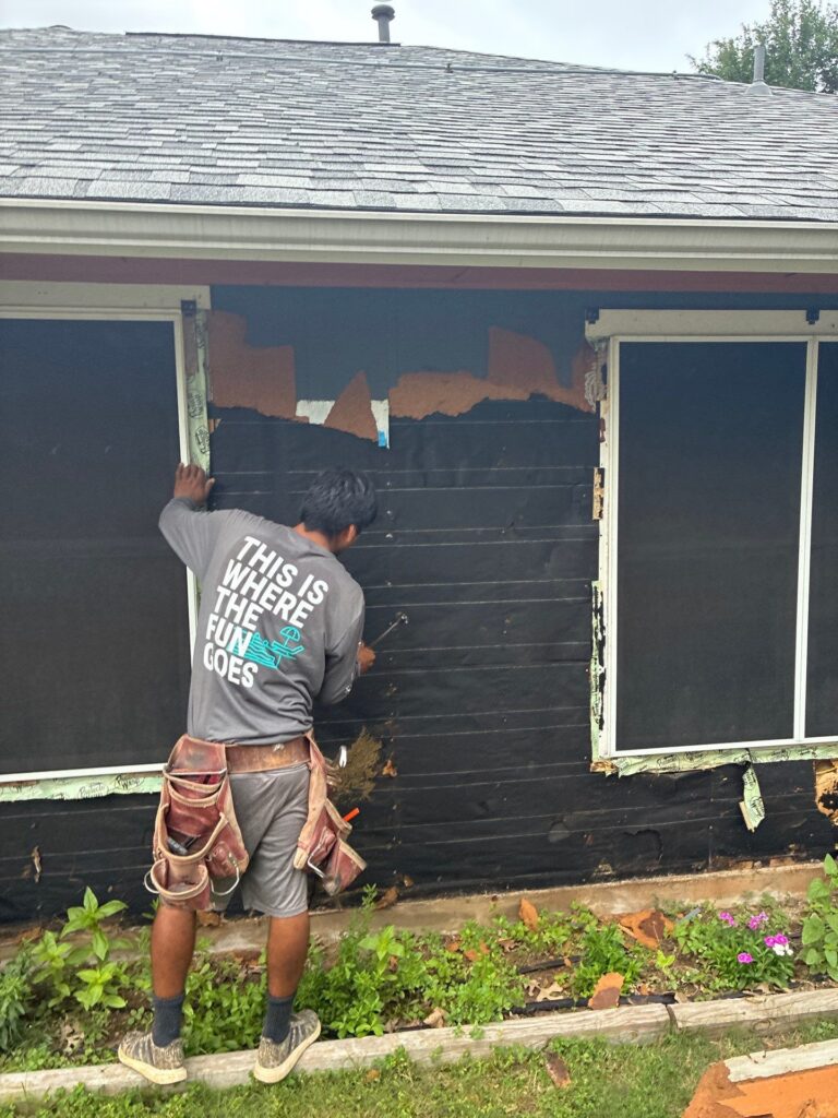 Construction worker in a tool belt removes black exterior siding from a house, exposing brown underlayment and a window frame; plants along the ground in the foreground.