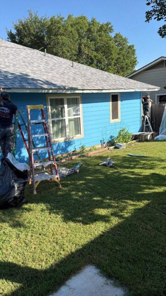 Blue, single-story house being renovated with workers on ladders in the front yard.