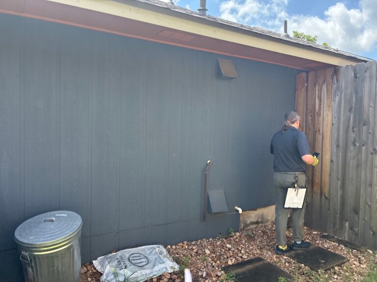 Person with a clipboard stands beside a dark blue wooden wall and a weathered wooden fence in a gravel yard.