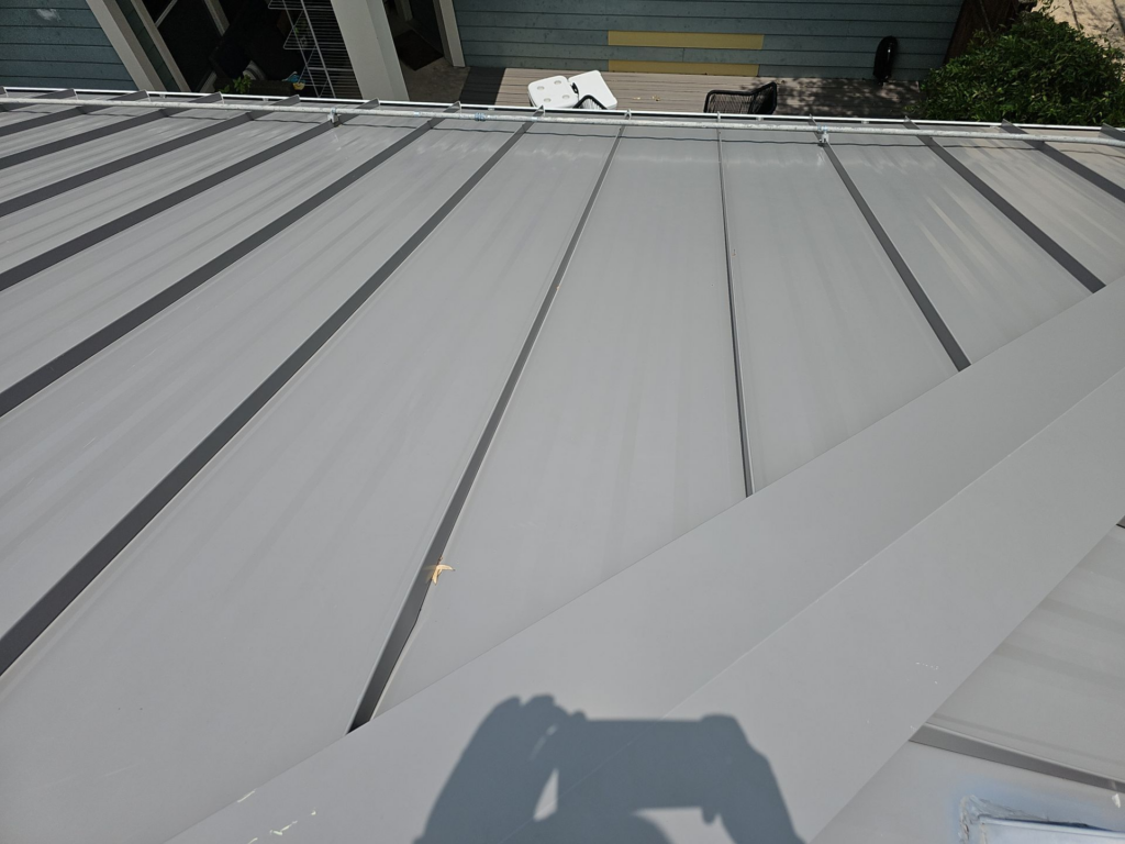 View from a gray metal standing-seam roof looking toward a small deck with a white chair and greenery in the background, photographer's shadow in foreground.