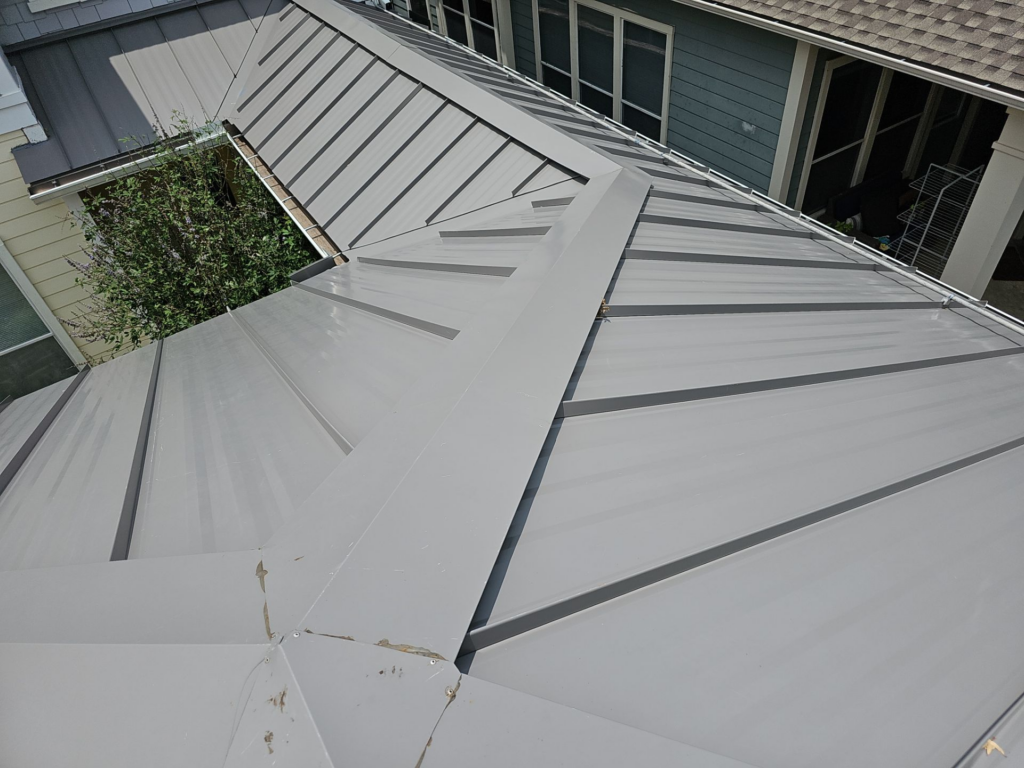 Aerial view of light gray metal standing-seam roof panels on a house, with a tree and neighboring walls nearby.