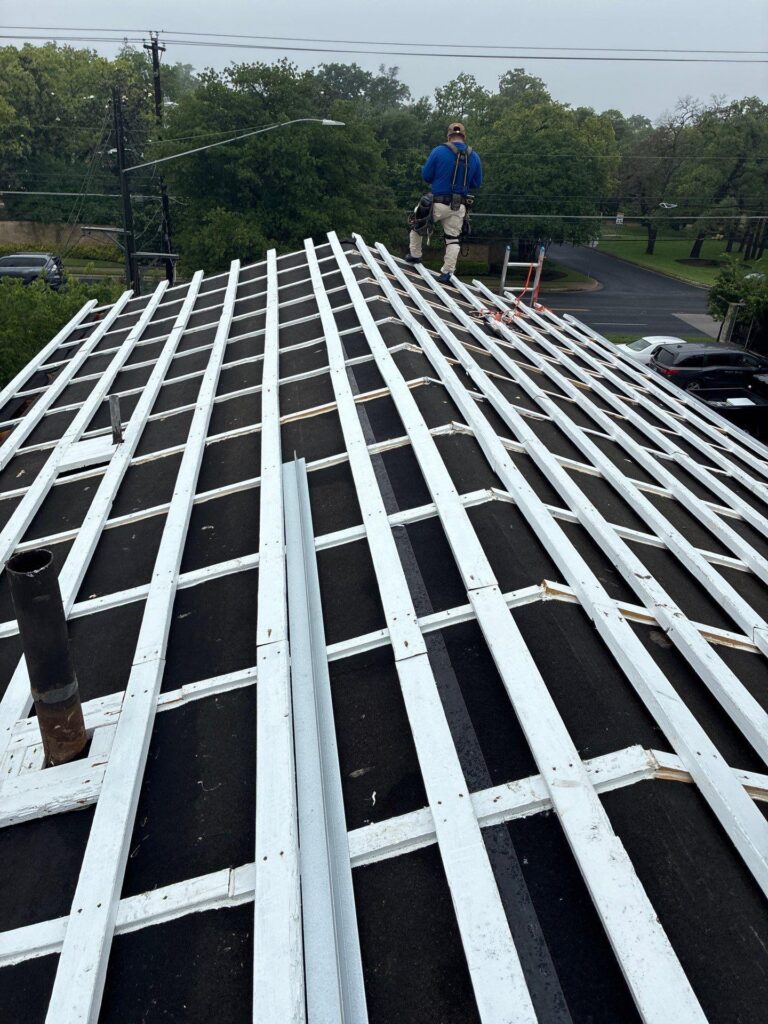 Worker in a blue shirt with safety harness on a pitched roof under construction, white rafter framing visible and debris around edge