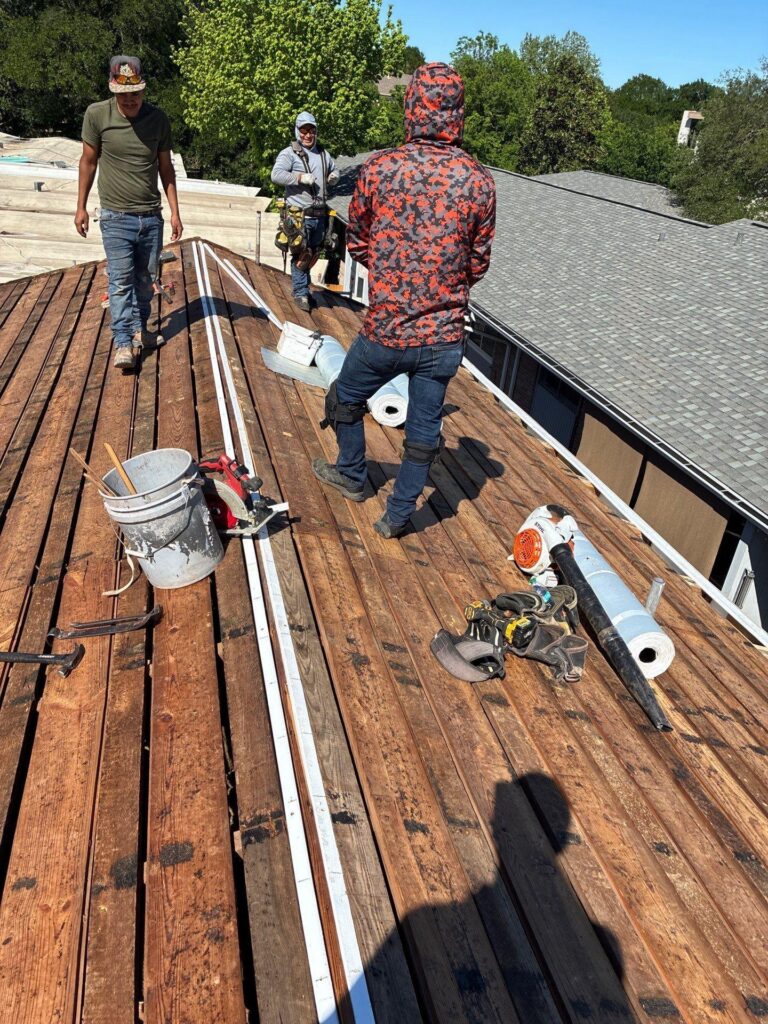 Three workers on a pitched wooden roof with tools, buckets, and roll of material during a roofing job.