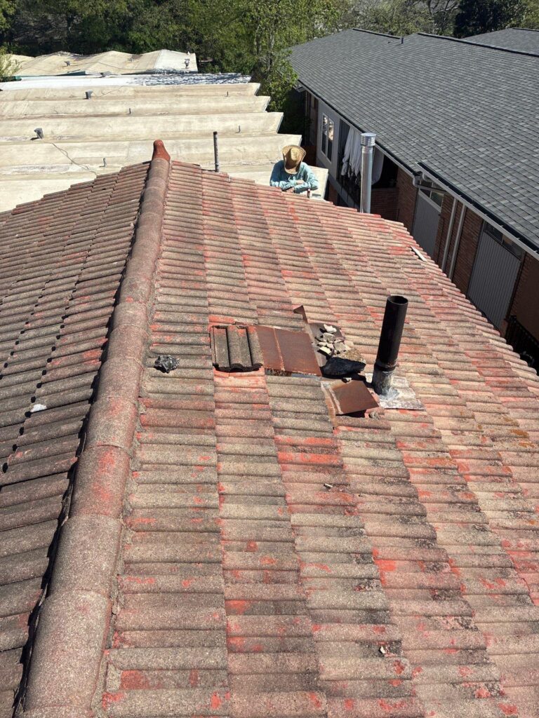 Worker in a hat standing on a red clay tile roof inspecting or repairing tiles, with debris nearby and neighboring roofs visible