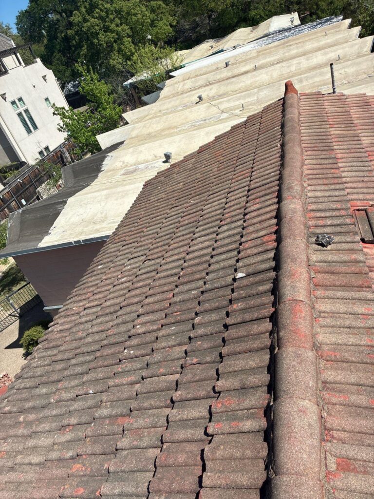 Aerial view of a red-tiled sloped roof in the foreground with beige flat roofs and green trees in the background.