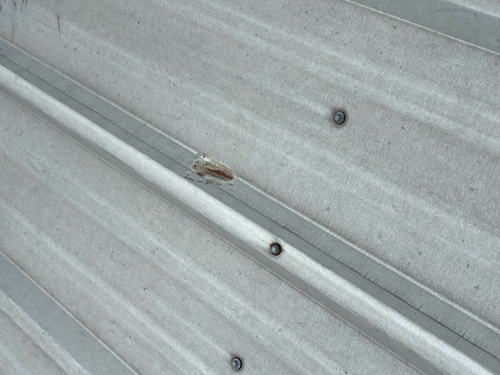 Close-up of a galvanized metal roof panel with screws along diagonal seams and a small rust spot nearby.