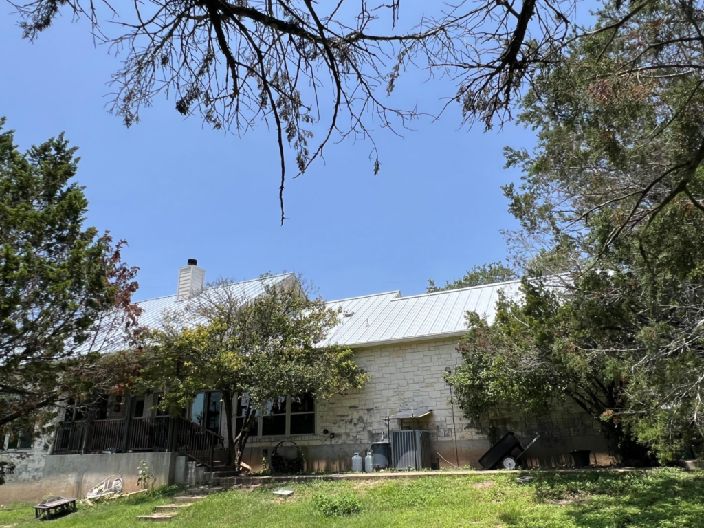 Backyard scene with a stone house and a metal roof, framed by leafy trees and a clear blue sky above.