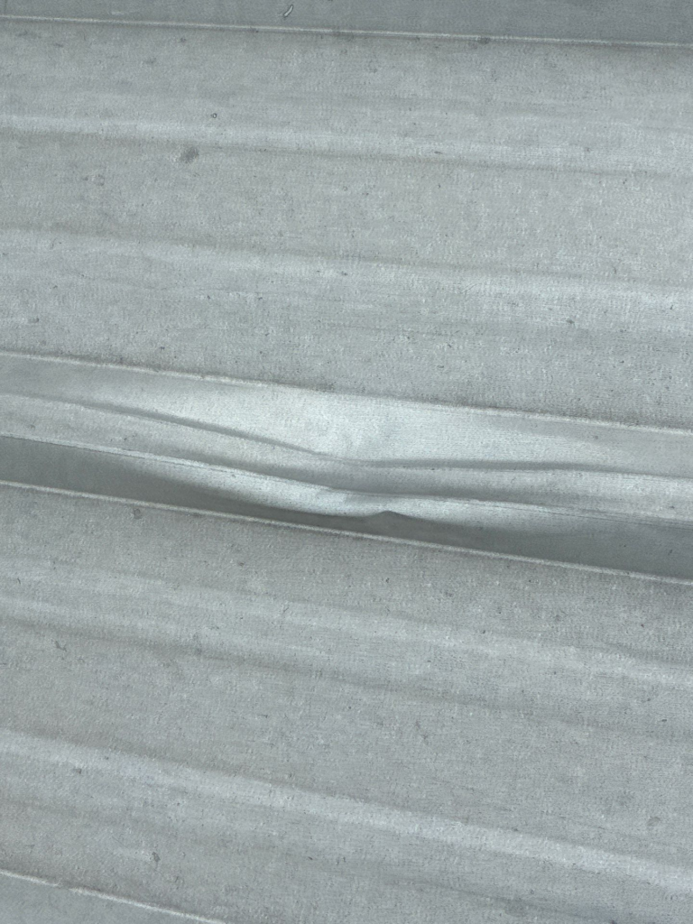 Wimberley_Before_4 - Driftwood Builders Roofing Close-up of a light-gray corrugated metal sheet with horizontal ridges and subtle weathering.