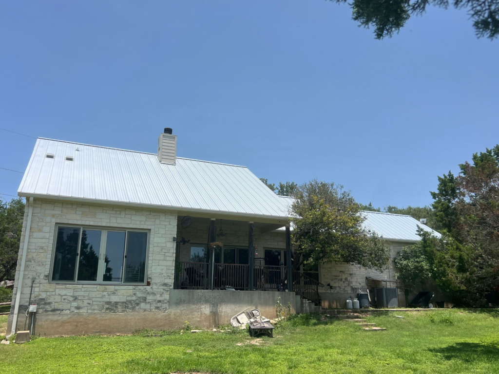 Front view of a stone house with a white metal roof, chimney, and a green lawnyard with trees nearby, under a blue sky, partial porch visible.