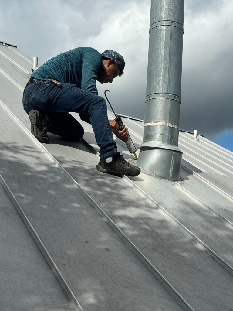 Roofer kneeling on a metal roof, sealing a seam around a vent pipe with a caulking gun.