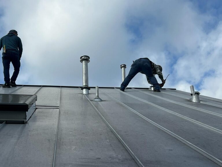 Two workers on a sloped metal roof, one standing near the edge and another bending with a hammer amid vents against a cloudy sky