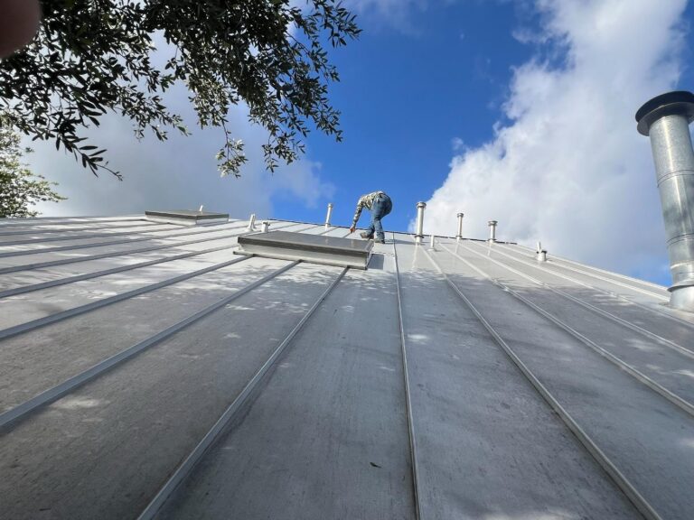 Worker on a sloped metal roof kneeling to install or secure a vent panel against a bright blue sky.