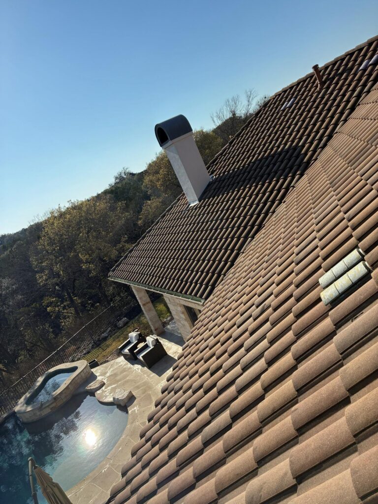 after_2 - Driftwood Builders Roofing Slanted brown tile roof with a white chimney vent, backyard pool area with lounge chairs and a hot tub, under a clear blue sky.