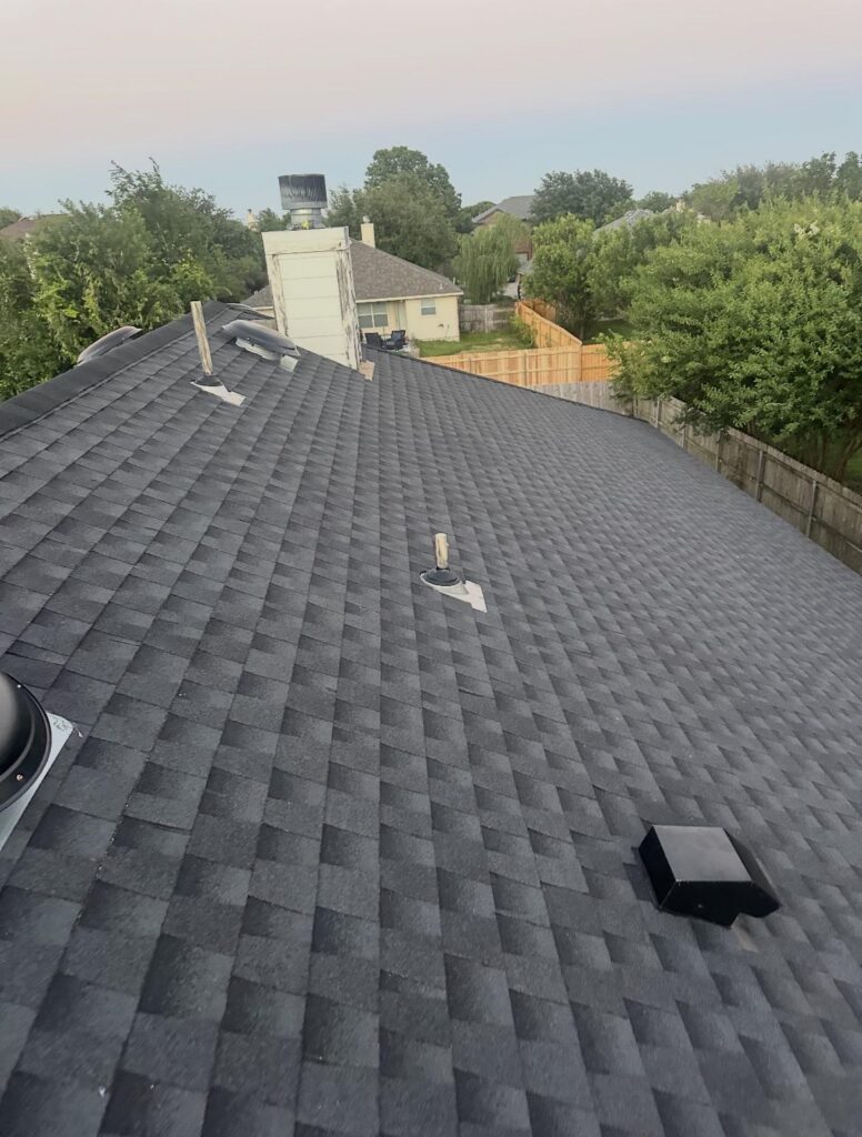 Aerial view of a dark gray shingled roof with roof vents and a chimney cap, neighboring houses and a fenced backyard in the distance under a pale sky.