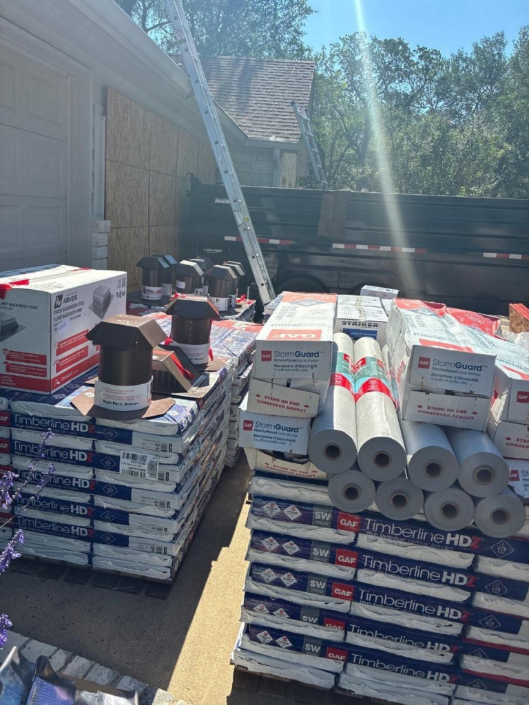 Stacks of roofing shingles and underlayment on pallets beside a house under construction, with ladders leaning on the wall and a dump trailer in the background.