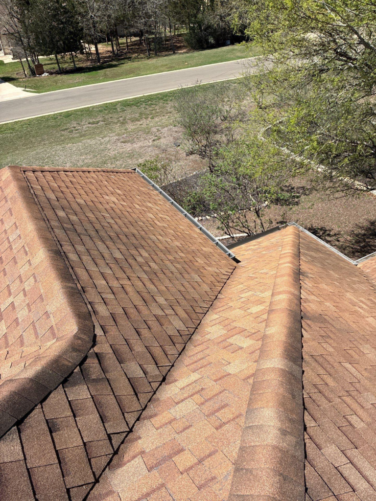 Aerial view of a brown shingle roof with intersecting ridges and surrounding yard and street beyond.
