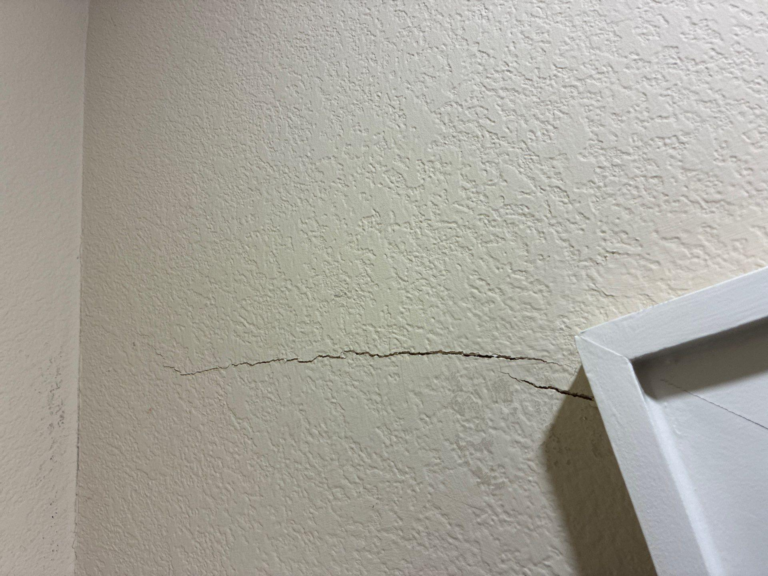 Beige textured interior wall with a long horizontal crack near mid-height; edge of a white cabinet visible on the right.