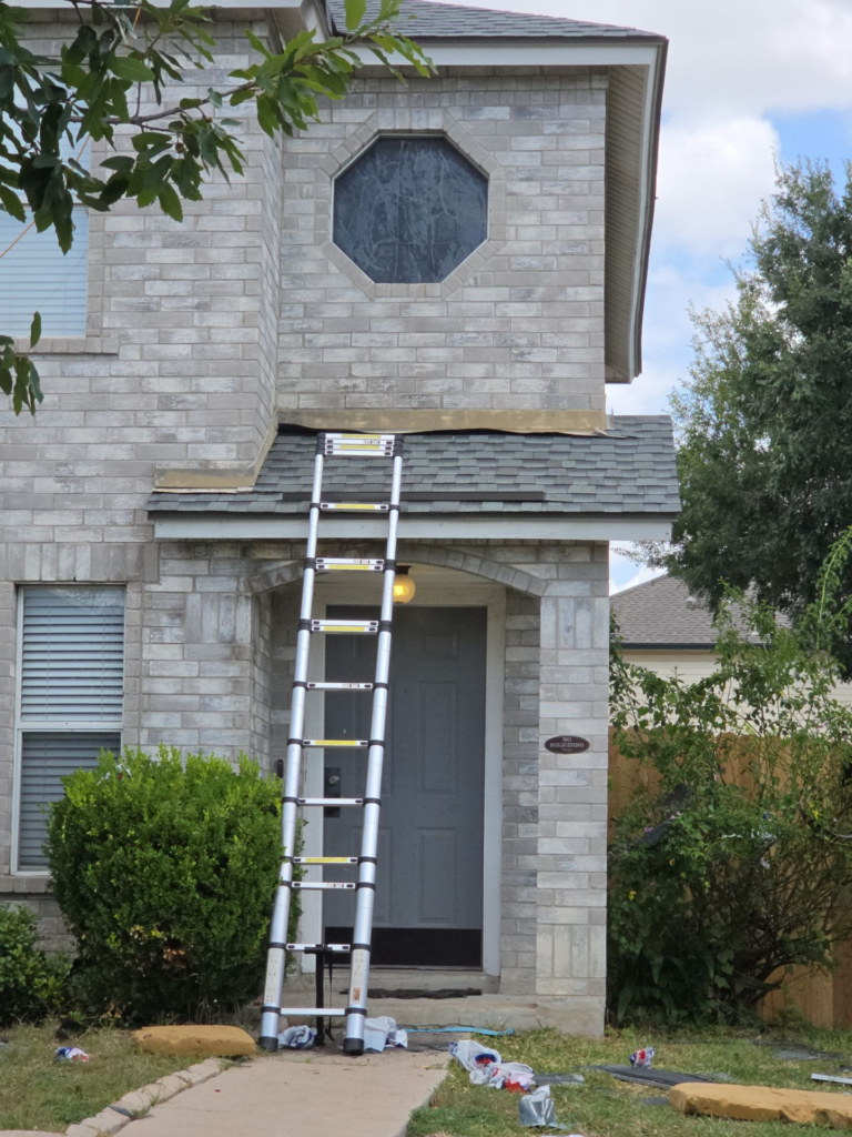 Aluminum ladder leaning against a two-story stone house under renovation, with an octagonal window above the entrance and debris on the yard.