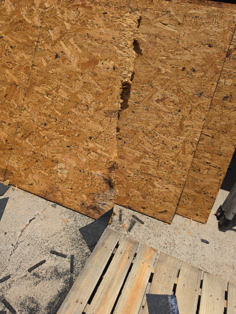 OSB plywood wall with a vertical split or gap, construction debris on the ground and a wooden pallet in the foreground.