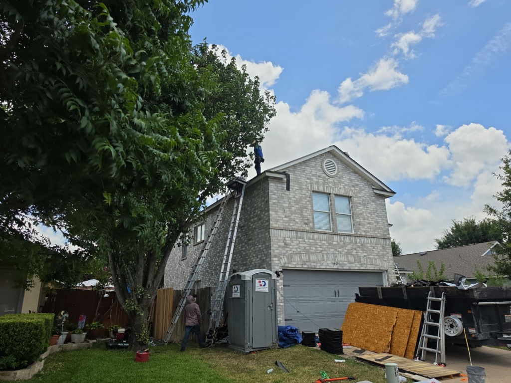 Worker on a ladder on the roof of a brick two-story house with construction materials and a portable toilet in the yard under a blue sky.