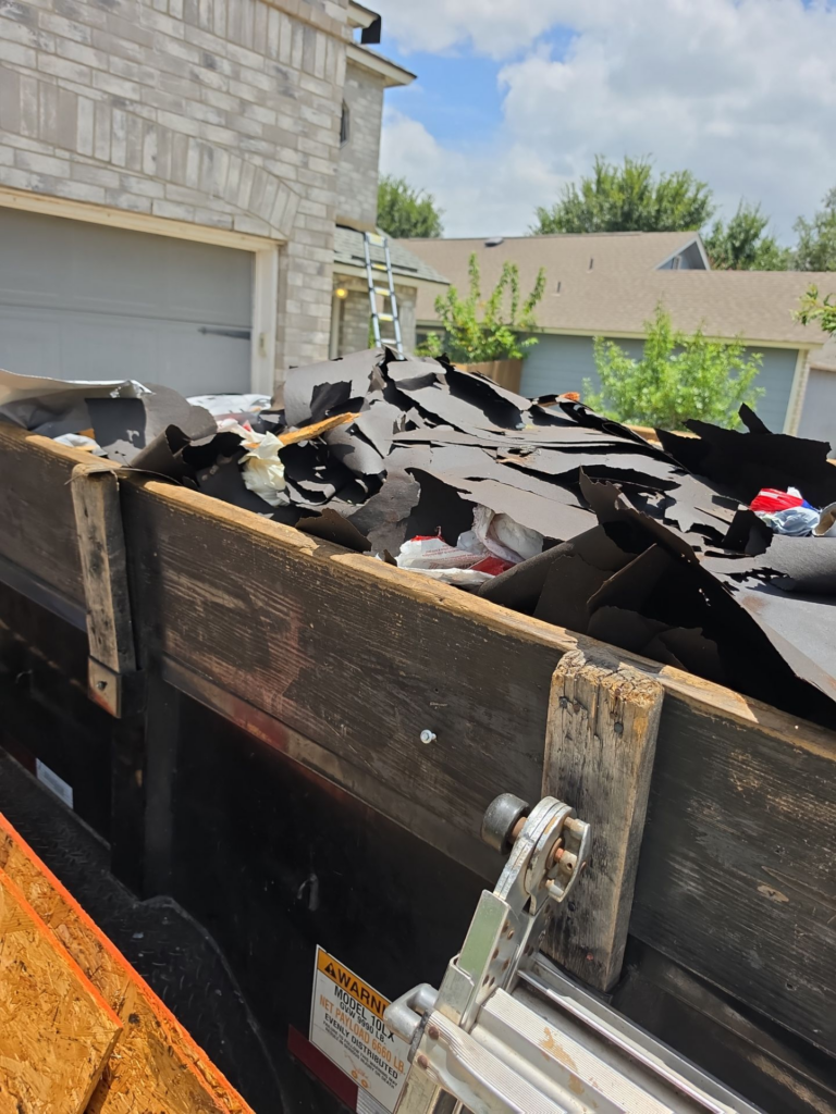Dumpster full of torn black roofing material and debris, with a ladder and suburban houses in the background.
