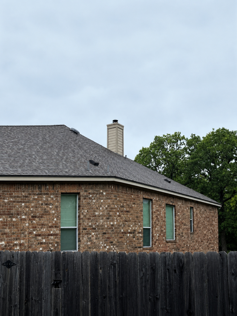 Brick house with a brown shingled roof and chimney, seen behind a tall dark wooden fence under a cloudy sky, with green-blind windows along the wall.